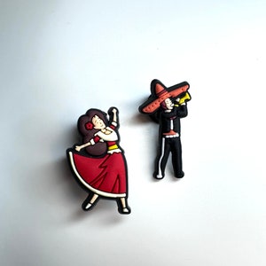 May include: A set of two colorful figurines depicting a man and woman in traditional Mexican attire. The man is wearing a black suit, a large sombrero, and playing a trumpet. The woman is wearing a red dress and a flower in her hair.