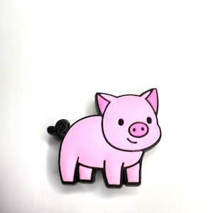 May include: A pink pig-shaped charm with black outlines and a curly tail. The pig has a smiling face with a pink snout and small black eyes. The charm is set against a white background.