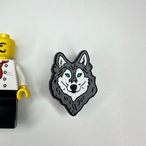 Puede incluir: Un amuleto de cabeza de lobo negro y gris con ojos verdes. El amuleto est&aacute; hecho de goma y tiene un dise&ntilde;o detallado.