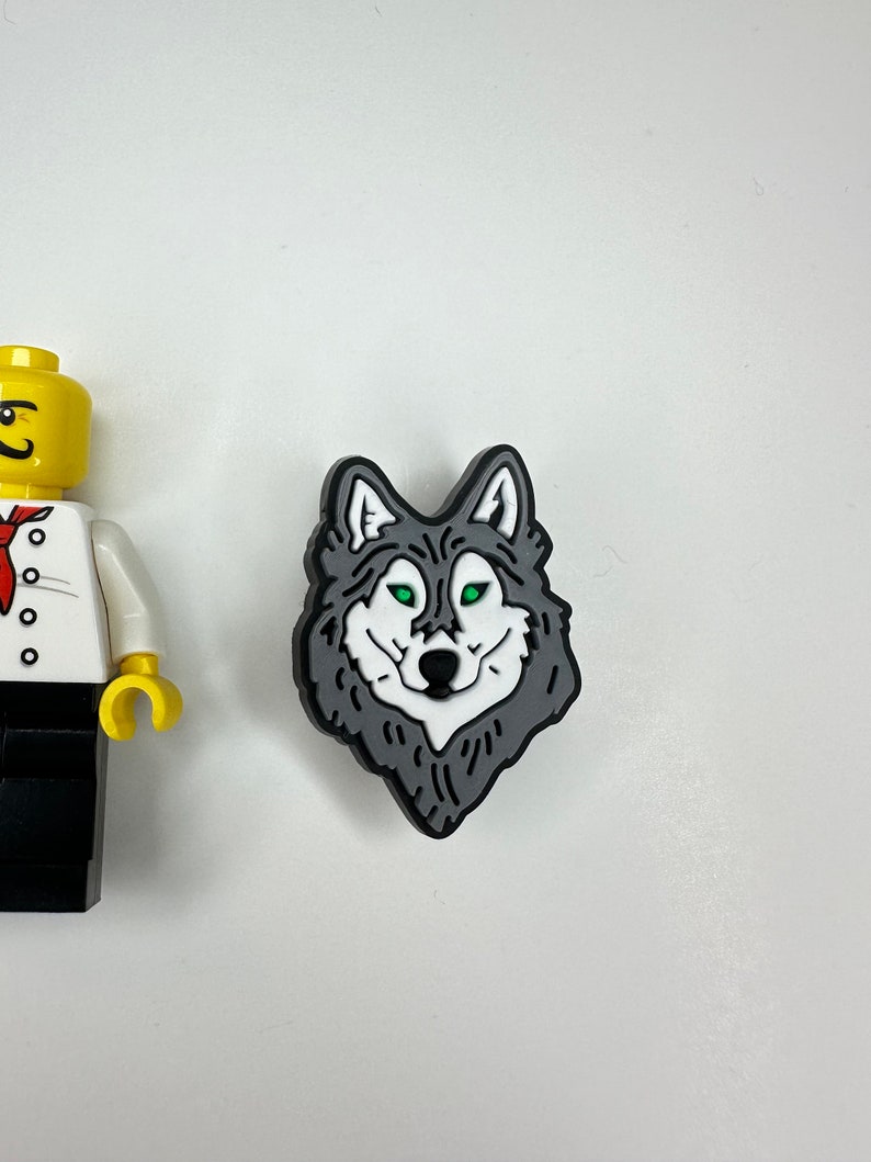 Puede incluir: Un amuleto de cabeza de lobo negro y gris con ojos verdes. El amuleto est&aacute; hecho de goma y tiene un dise&ntilde;o detallado.