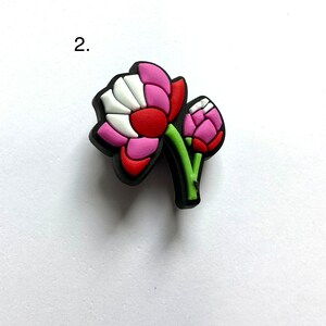 May include: A pink and red flower charm with a green stem and white accents. The charm is made of rubber and has a 3D design.