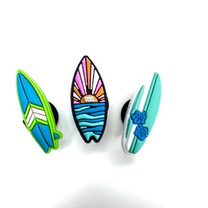 May include: Three colourful surfboard charms. One is blue and green with a white chevron design. The second has a sunset design with pink, orange, and blue colours. The third is white and teal with blue flower accents.