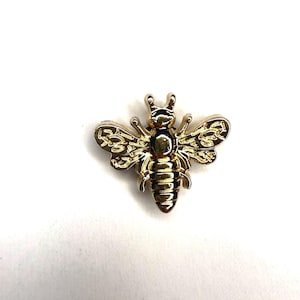 Può includere: Una spilla in metallo color oro a forma di ape con finitura lucida sulla testa e sull'addome, e dettagli incisi e testurizzati sulle ali. L'accessorio a forma di insetto presenta un design simmetrico con due antenne visibili sui lati.