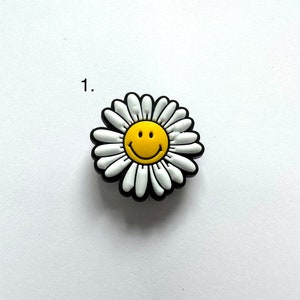 May include: A white daisy with a yellow smiley face in the center. The daisy is black outlined.
