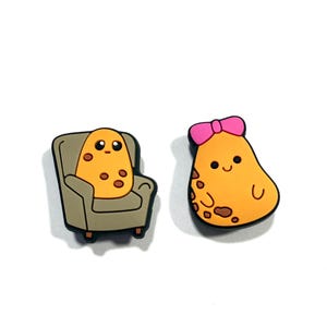 May include: Two cartoon potato-shaped charms. One is seated in a grey armchair, and the other has a pink bow. Both have brown spots and smiling faces. The charms are on a white background.