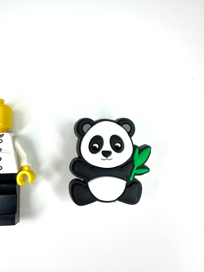 Puede incluir: Una figurita de panda blanco y negro sosteniendo un brote de bamb&uacute; verde. El panda est&aacute; sentado sobre una superficie blanca.