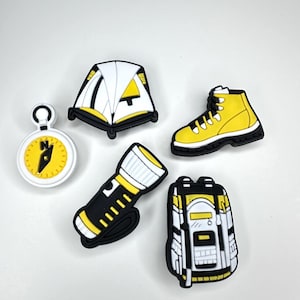 Puede incluir: Cinco amuletos de camping de color amarillo y negro. Los amuletos incluyen una tienda de campaña, una brújula, una linterna, una bota de senderismo y una mochila.