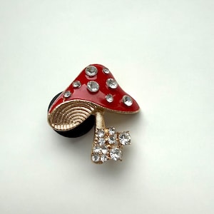 May include: A red mushroom-shaped charm with a gold stem and base, adorned with clear rhinestones. The mushroom cap is glossy red with embedded rhinestones. The charm is designed to attach to a shoe.