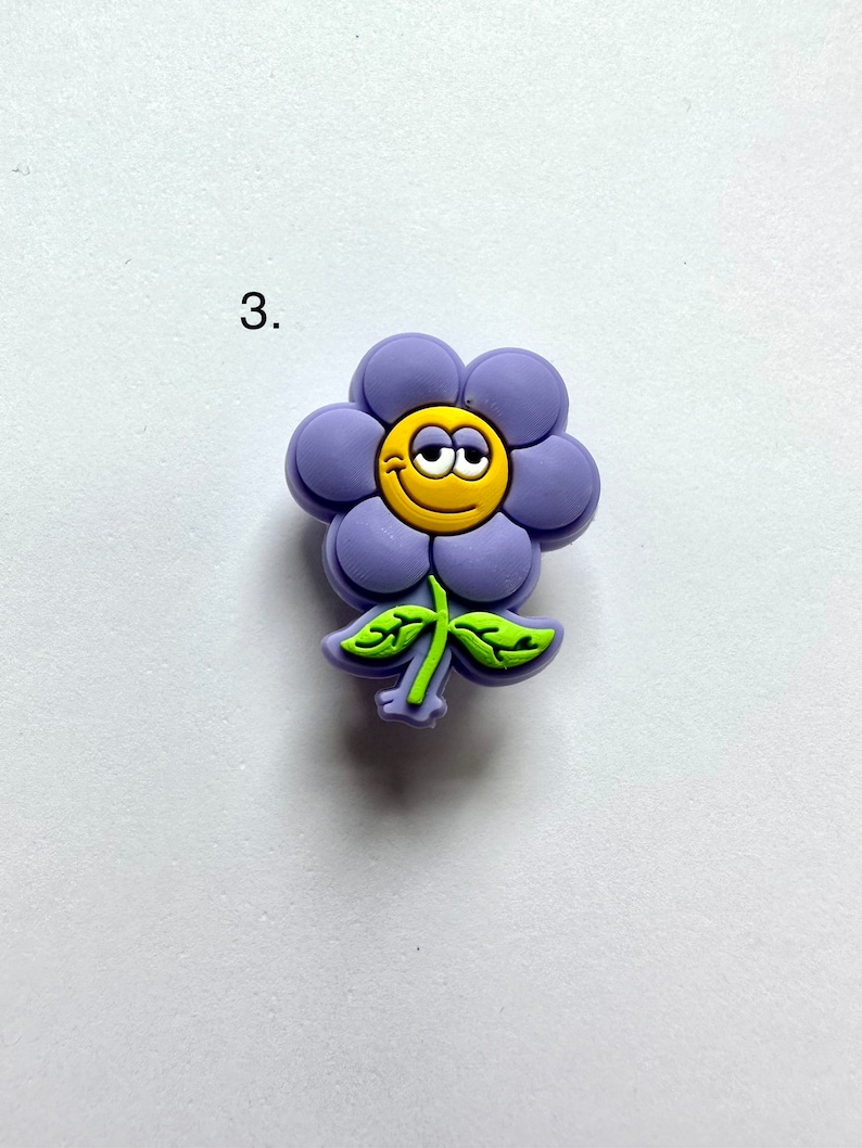 May include: Purple flower charm with a yellow smiley face and green leaves.