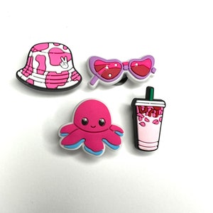 May include: A selection of shoe charms. Includes a pink and white cow print bucket hat, heart-shaped sunglasses, a pink octopus, and a pink drink charm with the word "Drink".