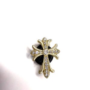 May include: A gold-coloured cross-shaped charm, adorned with clear, sparkling crystals. The cross is set on a small, round, black base. The design features a central crystal and smaller crystals along the arms of the cross.
