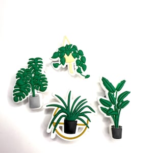 May include: Four plant-themed charms in green, white, and black. The charms include a hanging plant, a monstera, a spiky plant, and a plant with large leaves. Each plant is in a pot, with a white outline.