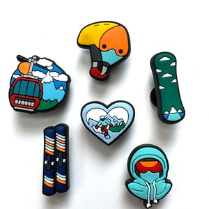 May include: A selection of ski and snowboard-themed charms in various colours. The collection includes a red and blue ski lift, a yellow and orange helmet, a green snowboard, a pair of skis, a heart-shaped charm with a skier, and a hooded figure.