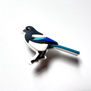 May include: A black and white magpie bird brooch with blue accents. The bird is perched on a white surface.