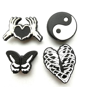 May include: Four black and white shoe charms. The charms include a heart held by skeleton hands, a yin-yang symbol, a butterfly, and a heart-shaped leaf with a skeleton ribcage design. These charms are likely for decorative purposes.