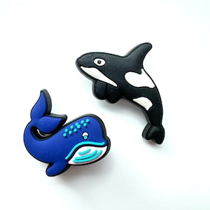 May include: Two blue and black whale shoe charms. One whale is a blue humpback whale with a white belly and the other is a black and white orca.