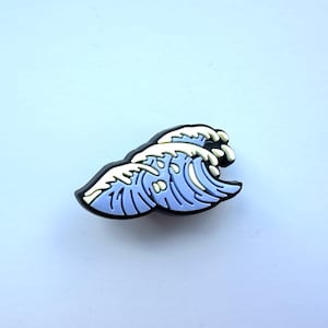 May include: A purple and white wave charm with a black outline. The charm is made of rubber and has a 3D design.
