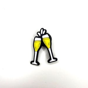 May include: A decorative pin featuring two champagne flutes clinking together. The flutes are filled with yellow liquid, outlined in black, and have white stems and bases. Above the glasses, there are three white shapes representing bubbles.