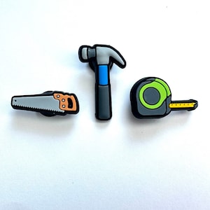 Pode incluir: Três amuletos de ferramentas coloridas: uma serra cinza, um martelo azul e cinza e uma fita métrica verde e preta.