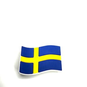 May include: A blue and yellow Swedish flag-shaped emblem with a white border. The flag features a yellow Nordic cross on a blue background. The emblem is likely a decorative item or accessory.