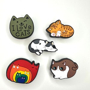 May include: Five colorful cat-themed shoe charms. One charm is green with the text "I love cats". Another charm is orange and white with a sleeping cat. A third charm is black and white with a cat lying on its belly. A fourth charm is brown and white with a cat sitting up. The fifth charm is red with a rainbow of cats stacked on top of each other.