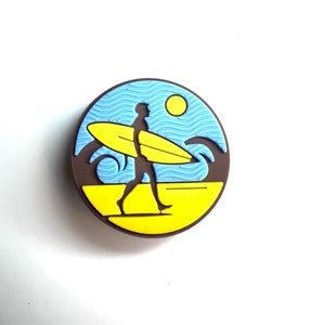 May include: A circular graphic depicting a surfer walking on a yellow beach, carrying a yellow surfboard. The background features blue waves, a blue sky, and a yellow sun. The graphic is framed by a brown border.