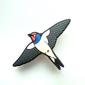 May include: A swallow bird figurine with a vibrant colour scheme. The bird has a red head, blue details, a white body, and grey wings. The swallow is a symbol of freedom and travel.
