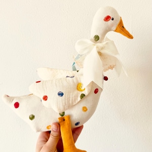 Peut inclure: Une peluche d'oie blanche faite à la main, ornée de pois colorés sur les ailes et le corps. L'oie a un bec et des pattes oranges, un nœud blanc et un point rouge et vert sur la tête. Le jouet est tenu par une main.