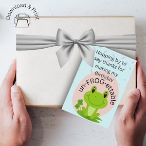 May include: A blue card with a green frog illustration and the text "Hopping by to say thanks for making my Birthday un-FROG-ettable". The card is tied with a silver ribbon to a white gift box.
