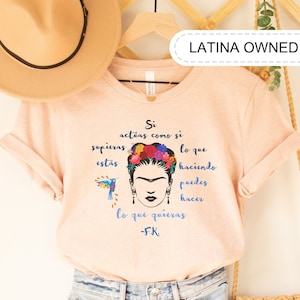Könnte beinhalten: Ein pinkfarbenes T-Shirt mit einem Grafikdruck von Frida Kahlo und einem spanischen Zitat, das lautet "Si actúas como si supieras, estás haciendo lo que puedes hacer lo que quieras -FK". Das Shirt hängt an einem Holzbügel mit einem braunen Hut und einer Strohtasche.