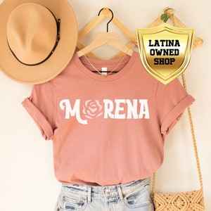 May include: A pink t-shirt with the word "MORENA" in white letters and a pink rose graphic. The shirt is on a hanger with a brown hat and a straw bag. The image also includes a gold shield with the text "LATINA OWNED SHOP".