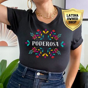 May include: A black t-shirt featuring a colourful floral design and the word "PODEROSA" in the centre. The shirt is being worn by a person with denim shorts. A gold shield with the text "LATINA OWNED SHOP" is in the top right corner of the image.
