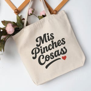 May include: Cream-colored canvas tote bag with the phrase "Mis Pinches Cosas" in black script, a small red heart, and a black underline. The bag is hanging on a wooden rack with pink flowers.