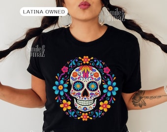 May include: Black t-shirt featuring a colorful sugar skull design. The skull is decorated with intricate floral patterns in shades of blue, yellow, orange, and pink, surrounded by a floral wreath. The text "LATINA OWNED" is in the top left corner.