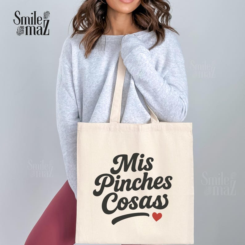 May include: Cream-colored tote bag with the phrase "Mis Pinches Cosas" in black script, a small red heart, and matching cream-colored handles. The bag is held by a person wearing a gray sweater.