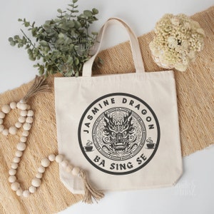 Puede incluir: Una bolsa de tela blanca con un diseño circular negro que presenta una cabeza de dragón y el texto "Jasmine Dragon Ba Sing Se".