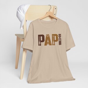 Op de afbeelding: Een beige t-shirt met het woord "PAPI" in bruin en goud gedrukt. De letters hebben een decoratief, gestileerd ontwerp. Een witte baseballpet rust op een houten stoel achter het shirt.