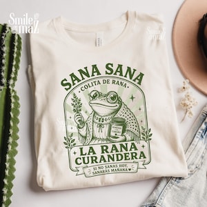 May include: A cream-colored t-shirt featuring a green graphic of a frog wearing glasses and a shawl. The frog holds a bundle of herbs and a jar labeled "Remedios con cariño". Text reads "Sana Sana Colita de Rana", "La Rana Curandera", and "Si no sanas hoy, sanarás mañana".
