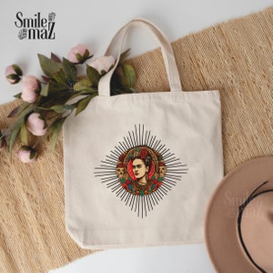 May include: A white canvas tote bag with a black and white illustration of Frida Kahlo surrounded by flowers and skulls. The illustration is in a circle with a sunburst design.