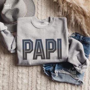 May include: A light gray sweatshirt with the word "PAPI" in large, dark blue block letters. The sweatshirt is displayed on a cream-colored knitted surface, with a gray hat and denim shorts visible in the background.