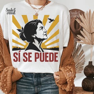 May include: White t-shirt featuring a black and gold graphic of a woman's profile, a bird, and the text "SÍ SE PUEDE" in red. The design includes a yellow sunburst background. The shirt is part of a clothing collection.