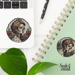 Puede incluir: Una pegatina con una ilustraci&oacute;n detallada de una mujer con maquillaje de calavera de az&uacute;car, adornada con flores y rodeada de elementos florales. La pegatina est&aacute; en un port&aacute;til y un cuaderno. Tambi&eacute;n se ve un bol&iacute;grafo.