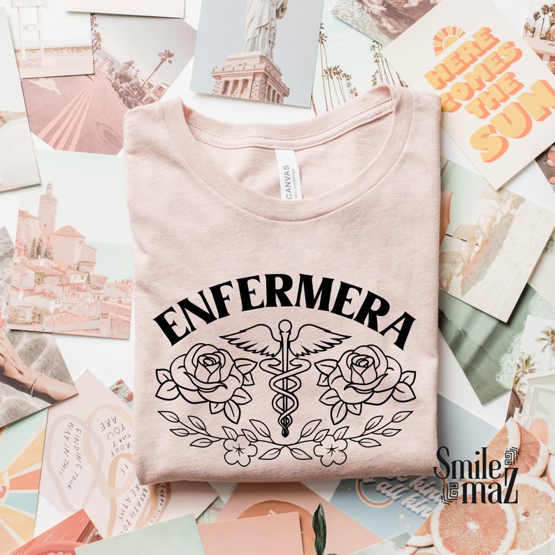 Pu&ograve; includere: Una maglietta rosa con un design grafico nero che dice "ENFERMERA" in uno stendardo circolare con un caduceo e rose.