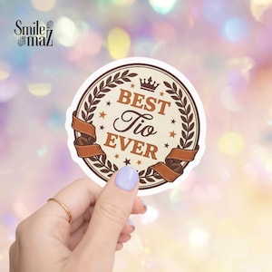 May include: A round sticker with the text "BEST Tio EVER" in brown and white. The text is surrounded by a brown laurel wreath and brown ribbons. Small stars are scattered around the text. The sticker has a white background.