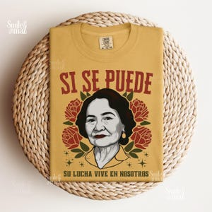 May include: Mustard yellow t-shirt with the phrase "SI SE PUEDE" in maroon lettering. The shirt features a portrait of a woman surrounded by red roses and the text "SU LUCHA VIVE EN NOSOTROS".