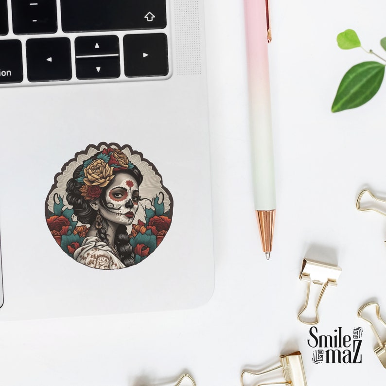 Puede incluir: Una pegatina redonda con una ilustraci&oacute;n detallada de una mujer con maquillaje de calavera de az&uacute;car, adornada con una corona floral y rodeada de rosas. La pegatina est&aacute; sobre un port&aacute;til, junto a un bol&iacute;grafo y material de oficina.