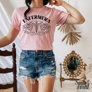 Pu&ograve; includere: Una maglietta rosa con la parola "ENFERMERA" in lettere nere, un simbolo di caduceo medico con ali e due rose con foglie. La maglietta &egrave; indossata da una persona con pantaloncini di jeans.