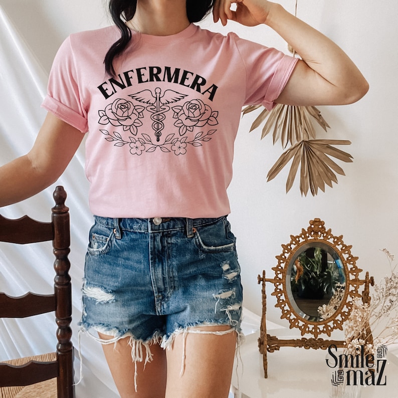 Pu&ograve; includere: Una maglietta rosa con la parola "ENFERMERA" in lettere nere, un simbolo di caduceo medico con ali e due rose con foglie. La maglietta &egrave; indossata da una persona con pantaloncini di jeans.