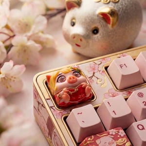 Puede incluir: Un teclado personalizado con teclas rosas y una tecla con temática de cerdo. Una hucha está en el fondo. El teclado tiene un marco dorado y un diseño floral. La tecla personalizada presenta un personaje de cerdo con un atuendo rojo.