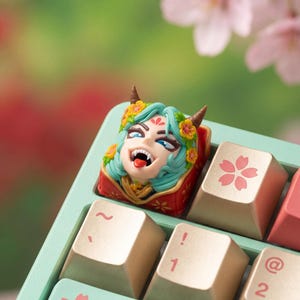 Può includere: Un keycap artigianale personalizzato con un design dettagliato e colorato di un demone femminile con capelli turchesi, corna e una corona floreale. Il keycap è rosso e oro, posto su una tastiera verde menta con tasti dorati.
